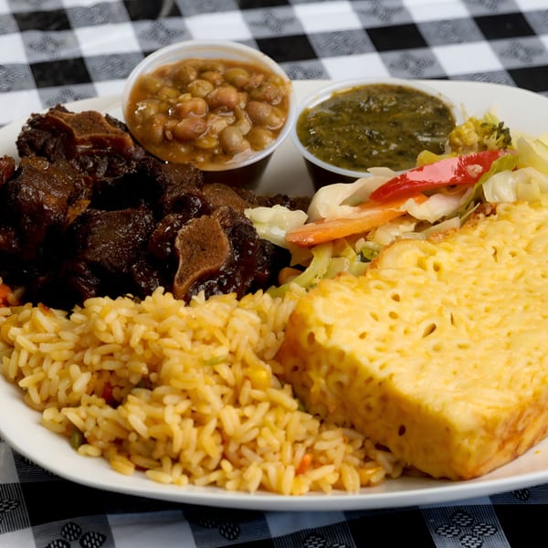 A Caribbean meal with oxtail, rice, mac and cheese, vegetables, and two sauces on a white plate.