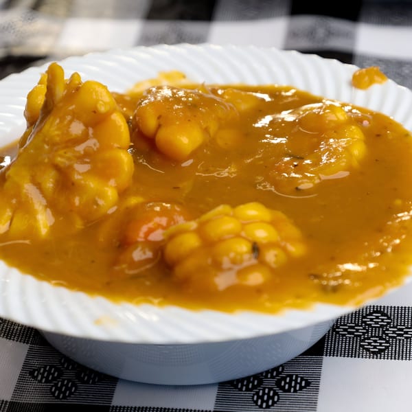 A white bowl filled with a vibrant orange Caribbean stew, featuring chunks of plantain and corn.