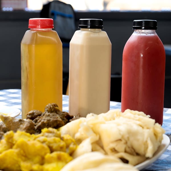 Three bottled Caribbean drinks, with yellow, cream, and red contents, alongside food on a checkered tablecloth.